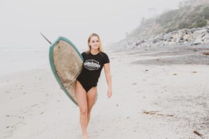 Blonde surfer girl walking on beach wearing black t-shirt holding a surfboard